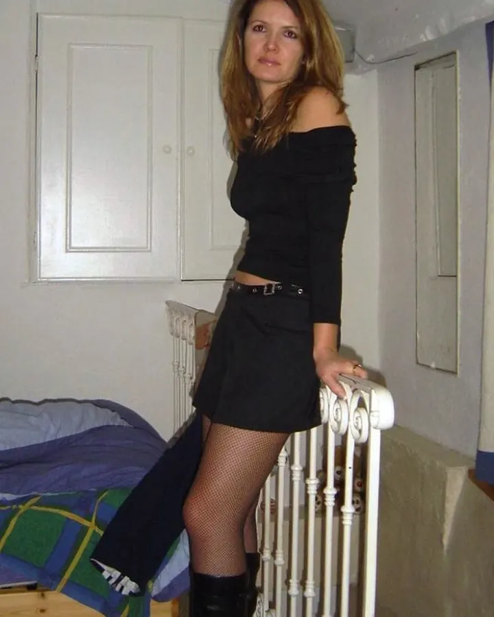 Femme aux cheveux blonds longs, en robe noire transparente, assise sur un lit blanc, les mains posées sur ses genoux.