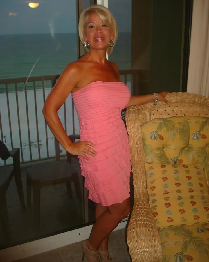Femme aux cheveux blonds courts, en robe rose sans bretelles, souriante, devant une baie vitrée donnant sur la mer.