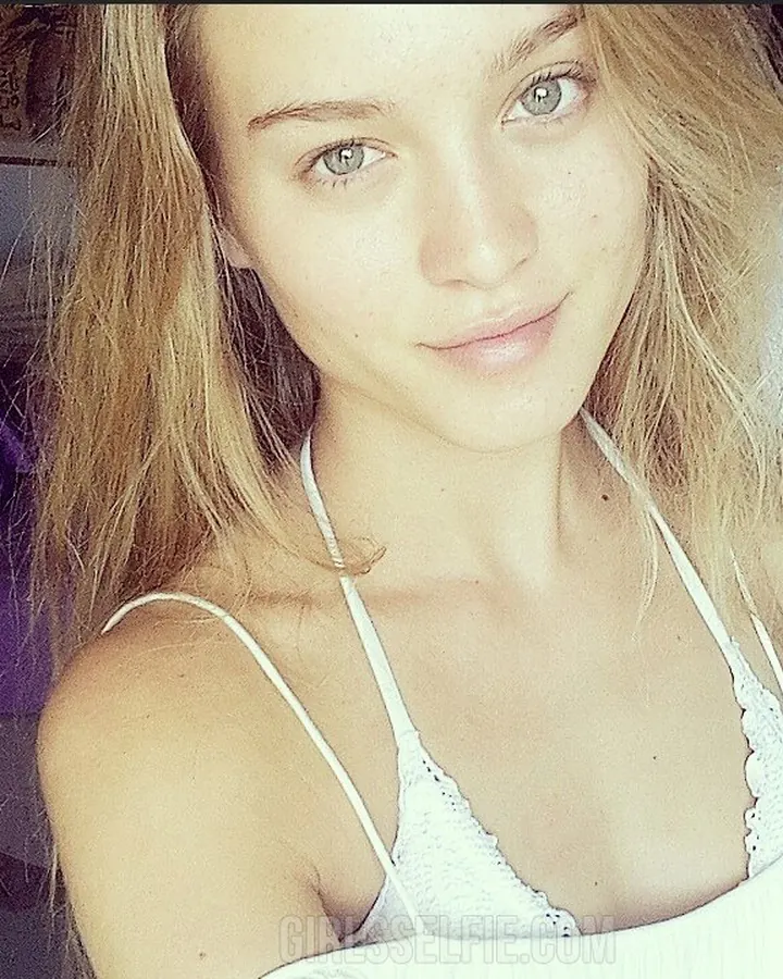 Jeune femme blonde, vue de profil, portant un haut blanc et un short beige, posant près d'un mur violet