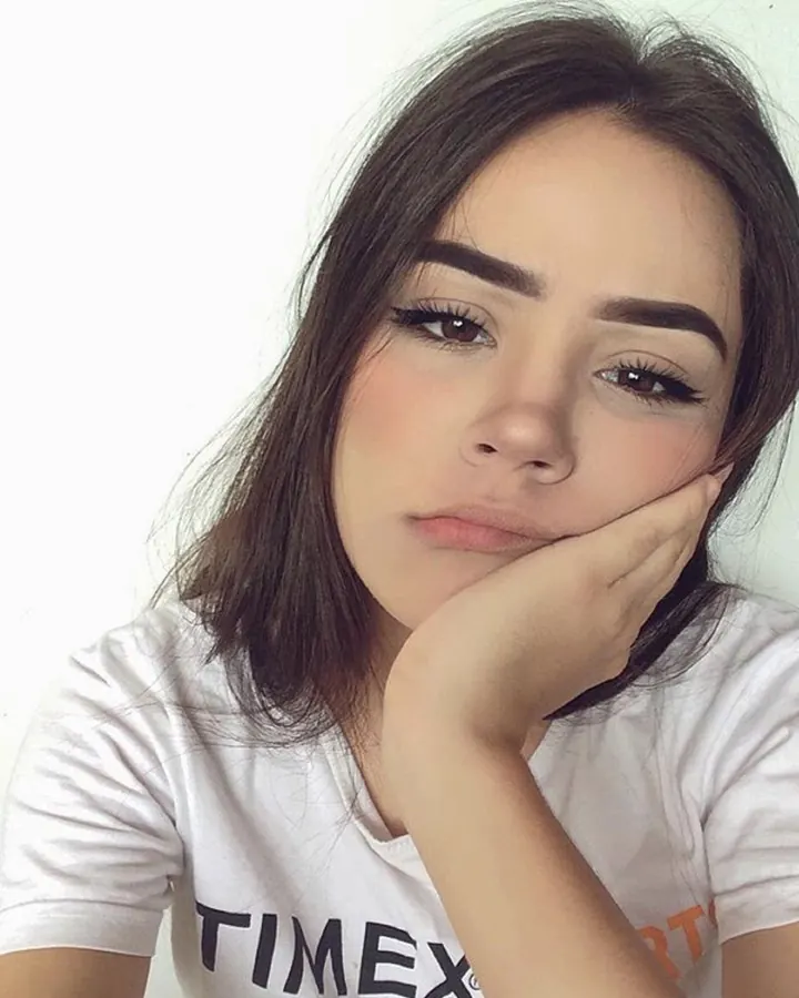 Jeune femme aux cheveux bruns, portant un t-shirt blanc avec le mot 'TIMEX', posant la main sous le menton