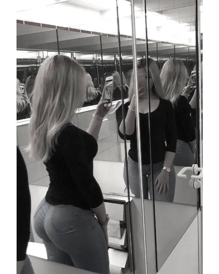 Jeune femme blonde en haut noir, prenant un selfie dans un miroir, vue de dos