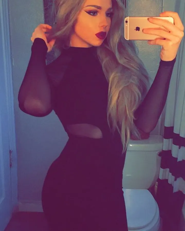 Jeune femme blonde en robe noire transparente, prenant un selfie devant un miroir dans une salle de bain