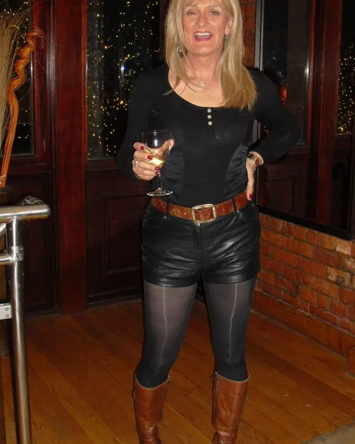 Femme aux cheveux blonds portant un haut noir, assise sur un canapé, tenant un verre, les jambes gainées de bas transparents.