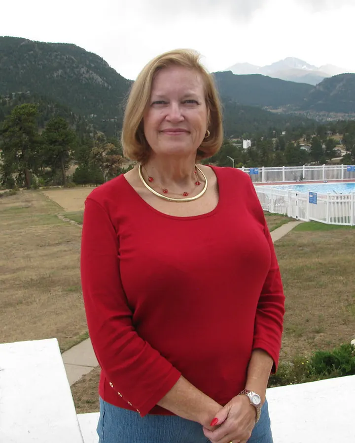 Femme souriante en pull rouge, debout sur une terrasse avec vue sur des montagnes et une piscine en arrière-plan.