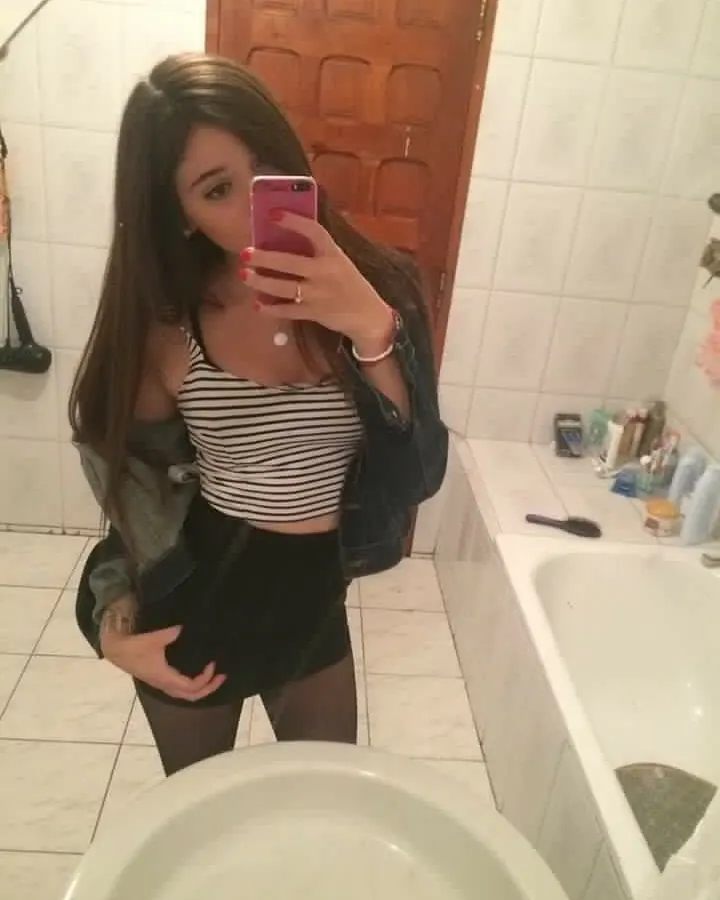 Jeune femme aux cheveux bruns, prenant un selfie dans une salle de bain, vêtue d'un haut rayé noir et blanc, jupe noire et collants foncés.