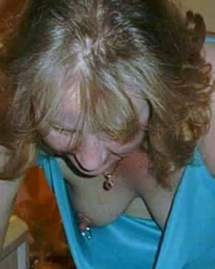 Femme en robe turquoise, assise sur un mur près d'une rivière, souriant à la caméra.