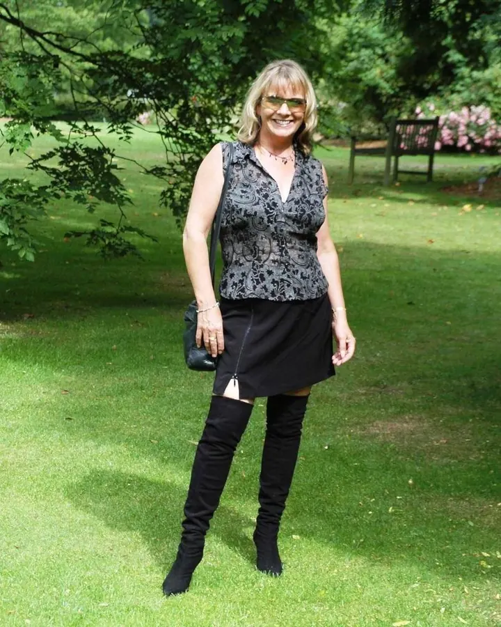 Femme souriante aux cheveux blonds, lunettes de soleil, robe noire et bottes hautes, debout sur une pelouse verte.