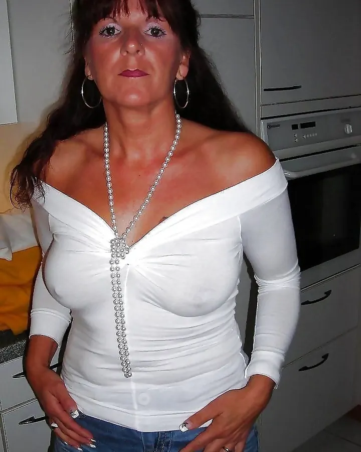 Femme en haut blanc, collier long, jeans, mains sur les hanches, dans une cuisine.
