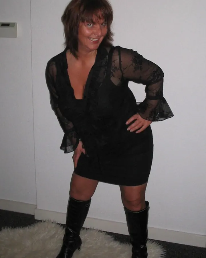 Femme en robe noire transparente, bottes hautes, souriante, posant sur une fourrure blanche.