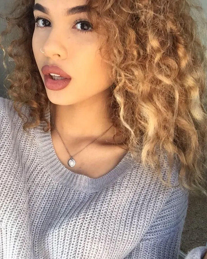 Jeune femme aux cheveux blonds bouclés, portant un pull gris clair, regardant l'appareil photo.