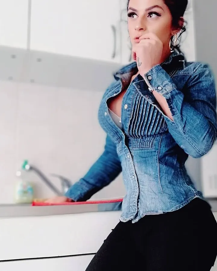 Femme aux cheveux noirs portant une chemise en jean bleu, tenant son menton, dans une cuisine.