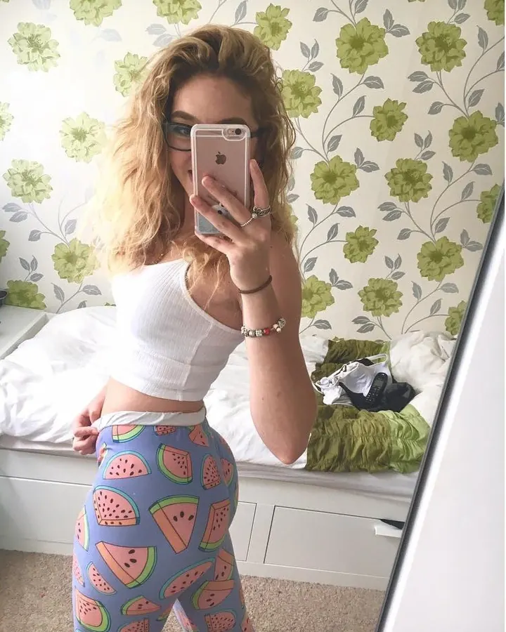 Femme blonde aux cheveux longs ondulés, portant un haut blanc, une jupe en cuir noir et des escarpins rouges, appuyée sur une voiture.