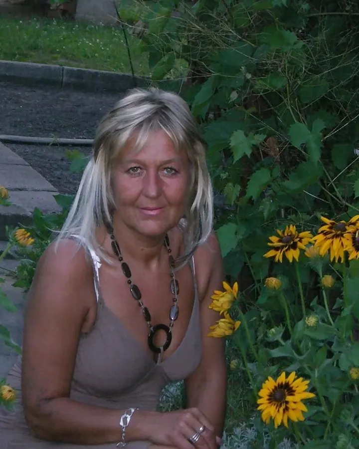 Femme blonde aux cheveux platine courts, portant un haut noir col roulé, assise sur un pouf noir, les jambes croisées, devant une vitrine en bois.