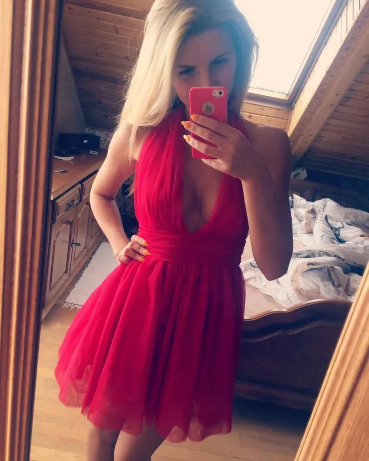 Femme blonde aux cheveux platine courts, portant un haut blanc, une jupe en cuir noir et des escarpins rouges, appuyée sur une voiture.