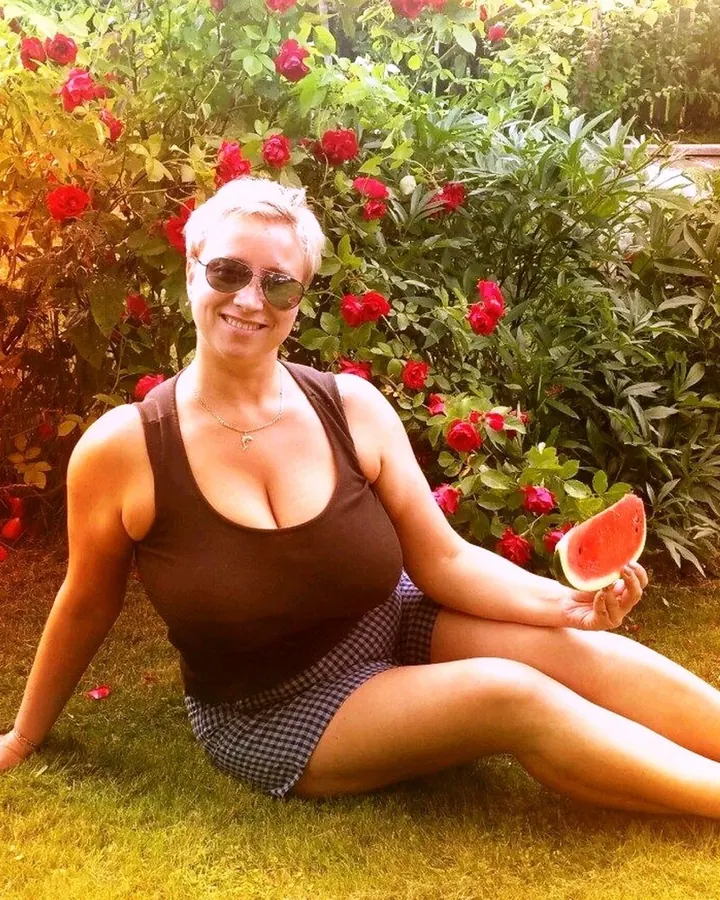 Femme aux cheveux gris, en haut noir, jupe à carreaux, posant sur l’herbe avec une pastèque, devant des roses rouges.