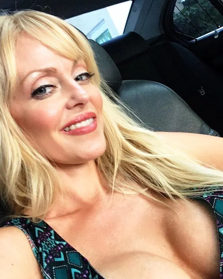 Femme blonde aux cheveux mi-longs, souriante, en haut imprimé, assise dans une voiture.