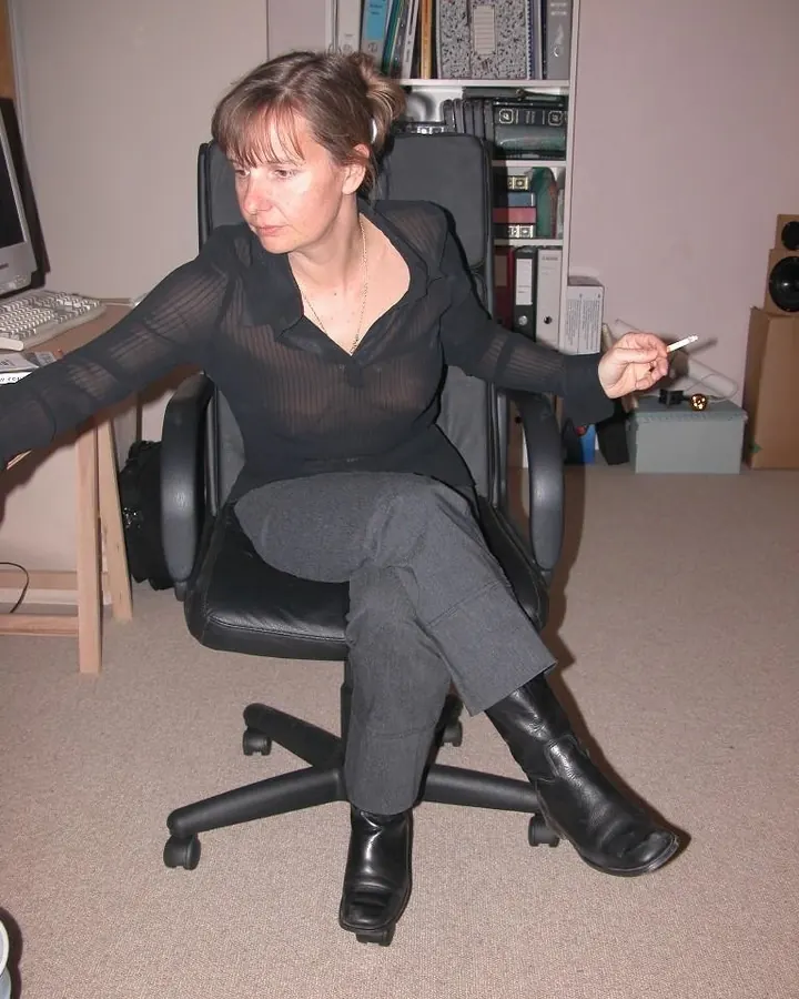 Femme aux cheveux châtains et yeux marrons en chemise noire transparente, assise sur une chaise de bureau.