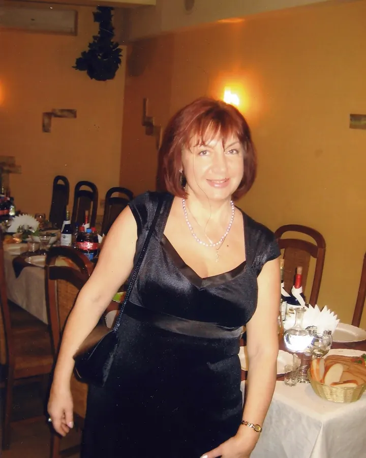 Femme aux cheveux roux souriant, vêtue d'une robe noire longue, portant un collier de perles, dans une salle à manger élégante.
