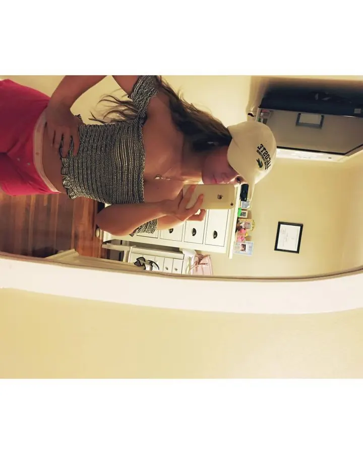 Jeune femme aux cheveux blonds prenant un selfie devant un miroir, portant un haut rayé et un short rose, coiffée d'une casquette blanche.