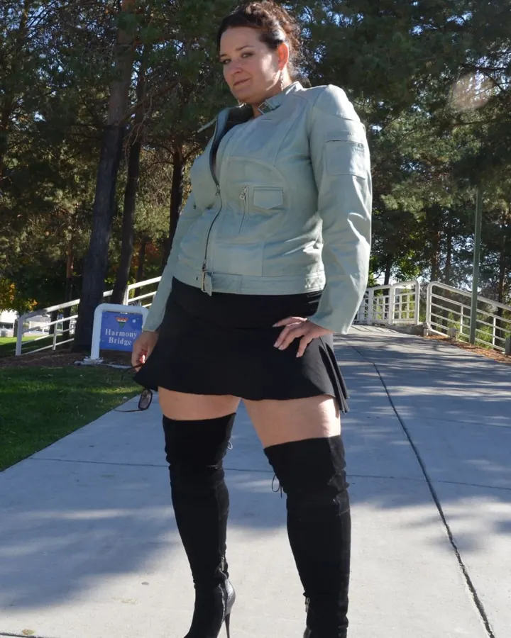 Femme aux cheveux châtains posant dehors, vêtue d'une veste grise, d'une jupe noire courte et de bottes noires au-dessus du genou.