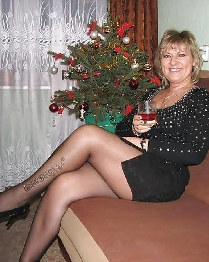 Femme aux cheveux blonds courts, en robe noire courte, assise sur un canapé, tenant un verre de vin rouge, devant un sapin décoré.