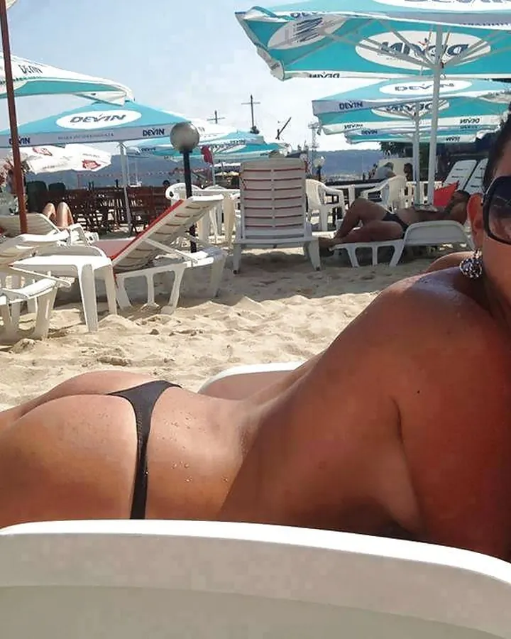 Femme latino aux cheveux bruns-noirs, vêtue d'un maillot de bain noir, allongée sur une chaise longue au bord de la plage, sous des parasols bleus.