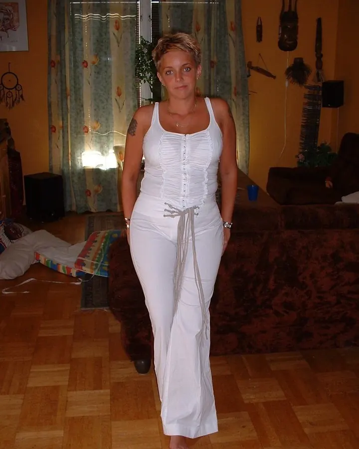 Jeune femme blonde aux yeux bleus, vêtue d'un ensemble blanc corseté, debout dans un salon avec des rideaux floraux et des objets décoratifs autour.