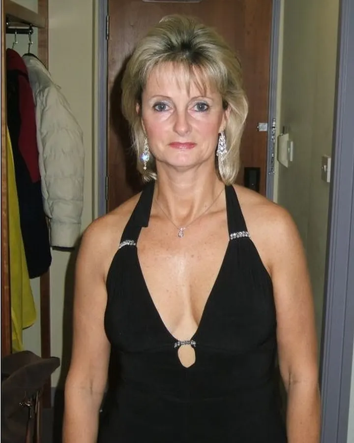 Femme blonde aux yeux gris, vêtue d'une robe noire à col halter, d'un collier et de boucles d'oreilles, debout dans un couloir, regardant droit devant.