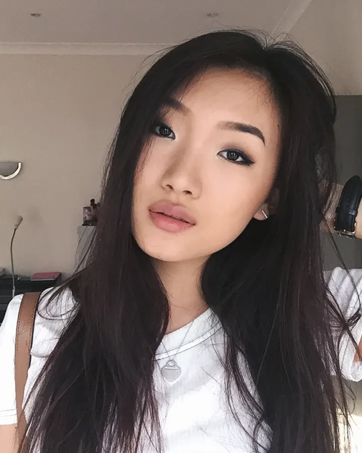 Femme asiatique aux cheveux bruns-noirs, yeux noirs, vêtue d'un haut noir, d'un short blanc et de collants gris, en train de marcher dans un couloir, les jambes gainées et les chevilles montées.