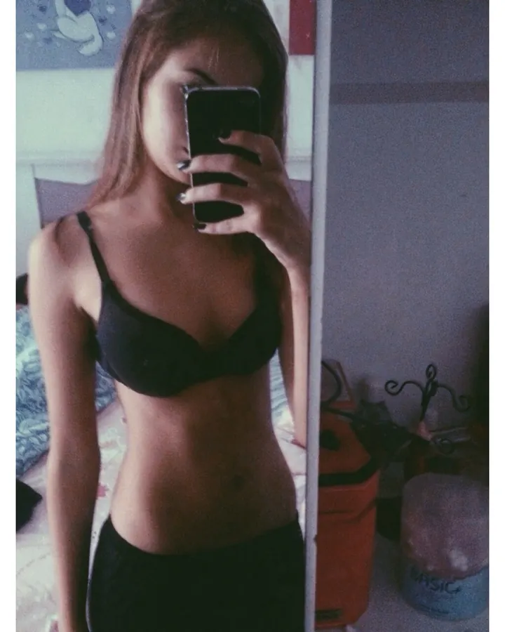 Femme aux cheveux bruns portant un soutien-gorge noir, se prenant en photo dans un miroir, tenant un téléphone noir, dans une chambre.