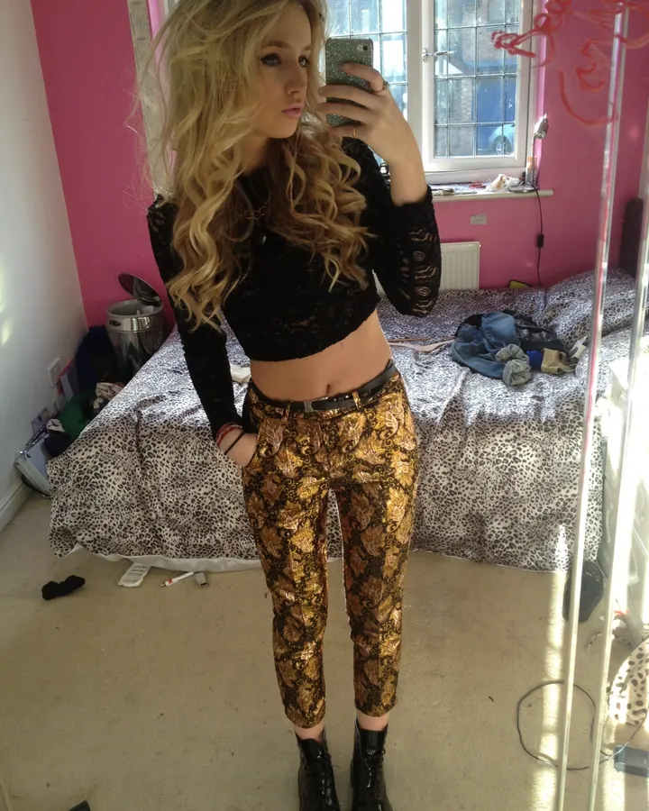 Jeune femme blonde aux cheveux ondulés portant un haut noir ajouré et un pantalon doré, prenant un selfie dans une chambre rose.