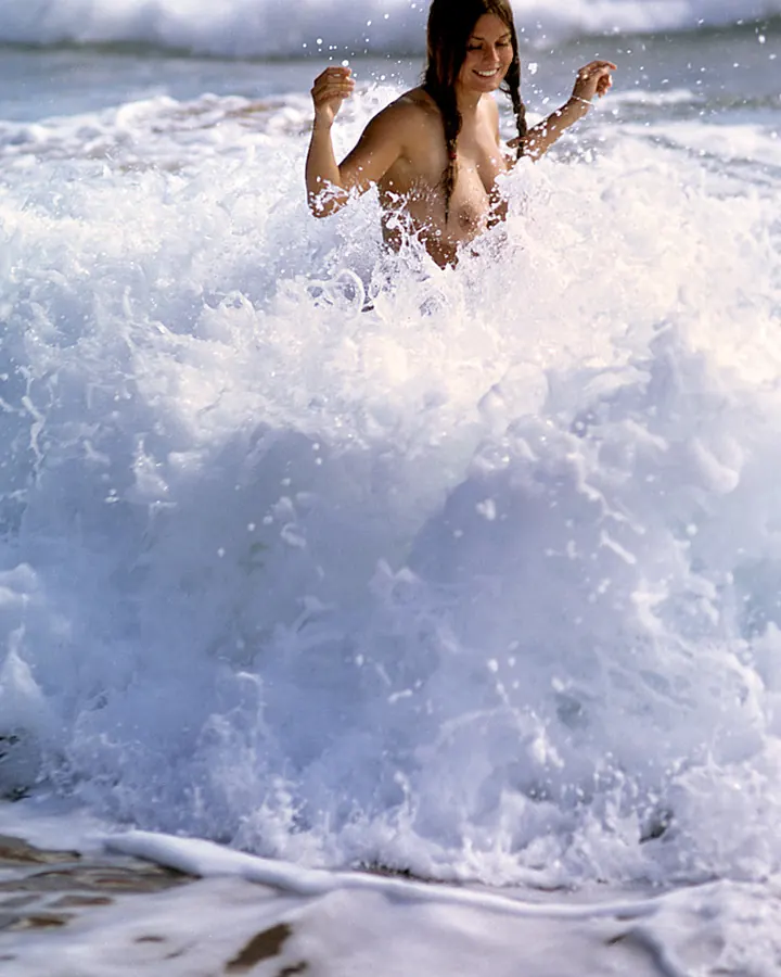Femme aux cheveux bruns, nue, dans les vagues de la mer, souriant.