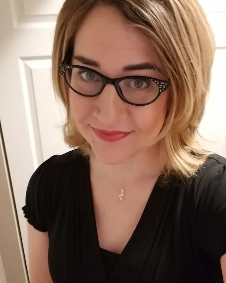 Femme aux cheveux blonds, lunettes, en haut noir, souriant légèrement devant une porte blanche.