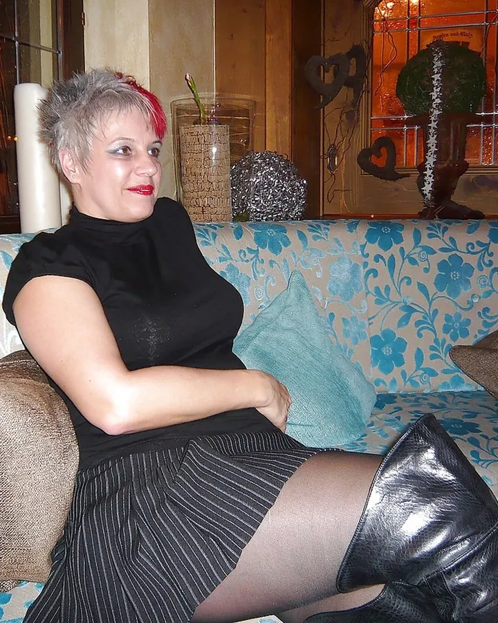 Femme blonde en haut noir, assise sur un canapé, regardant vers la caméra, main sur la poitrine.
