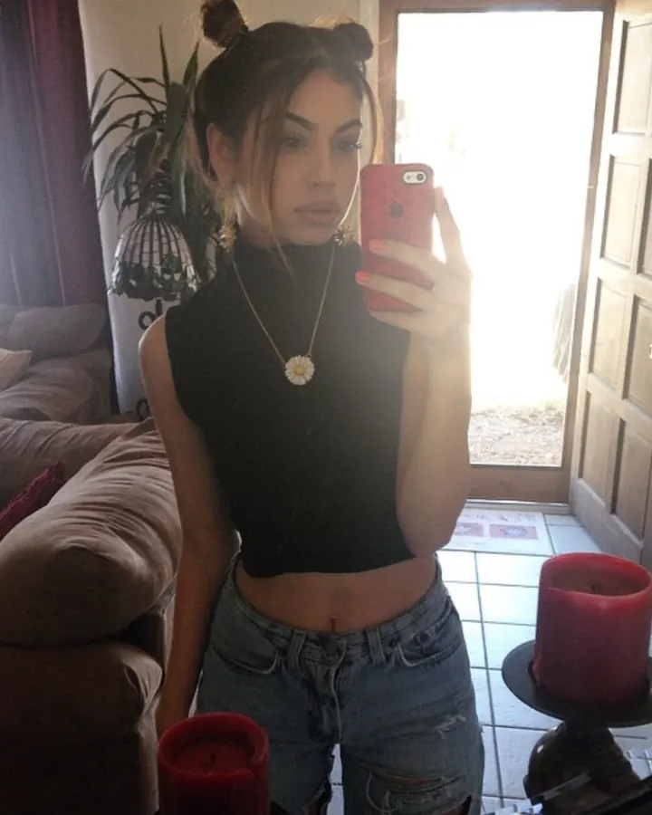 Femme brune en haut noir et short court, vue de dos, appuyée sur une voiture noire, talons hauts.