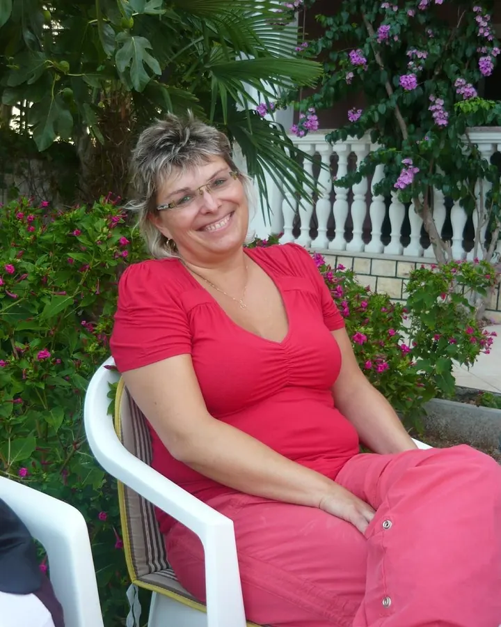 Femme blonde en tenue rose, assise sur une chaise, souriante, entourée de fleurs.