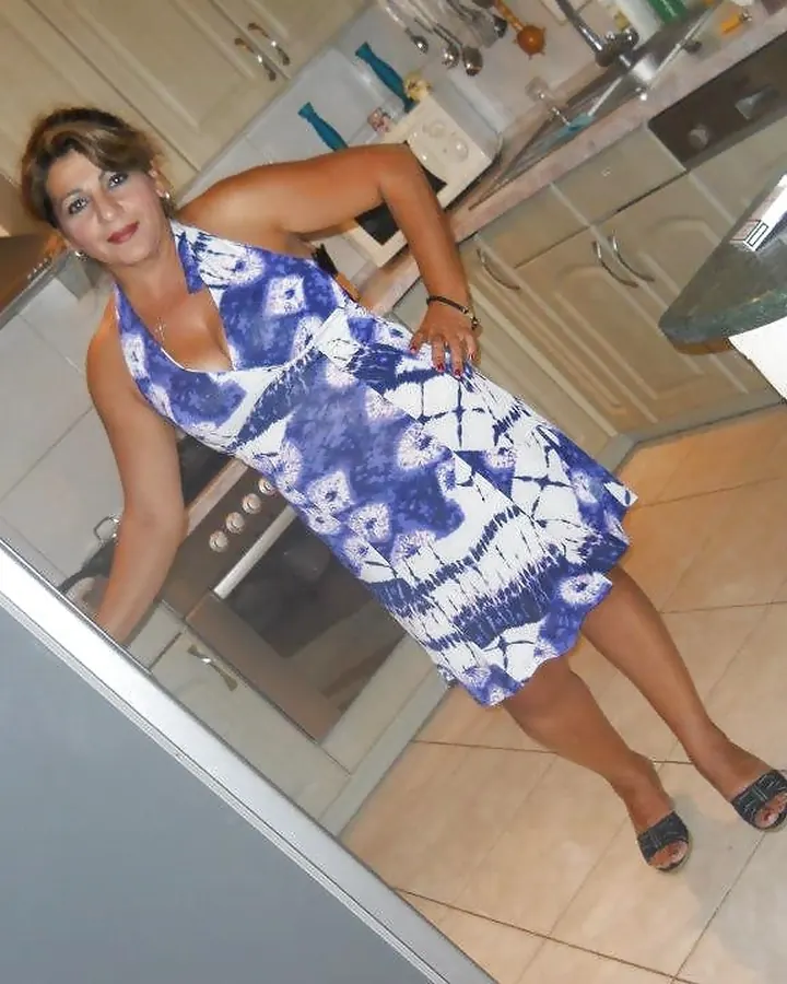 Femme aux cheveux bruns, en robe blanche, posant dans un salon avec un fond de peinture florale.