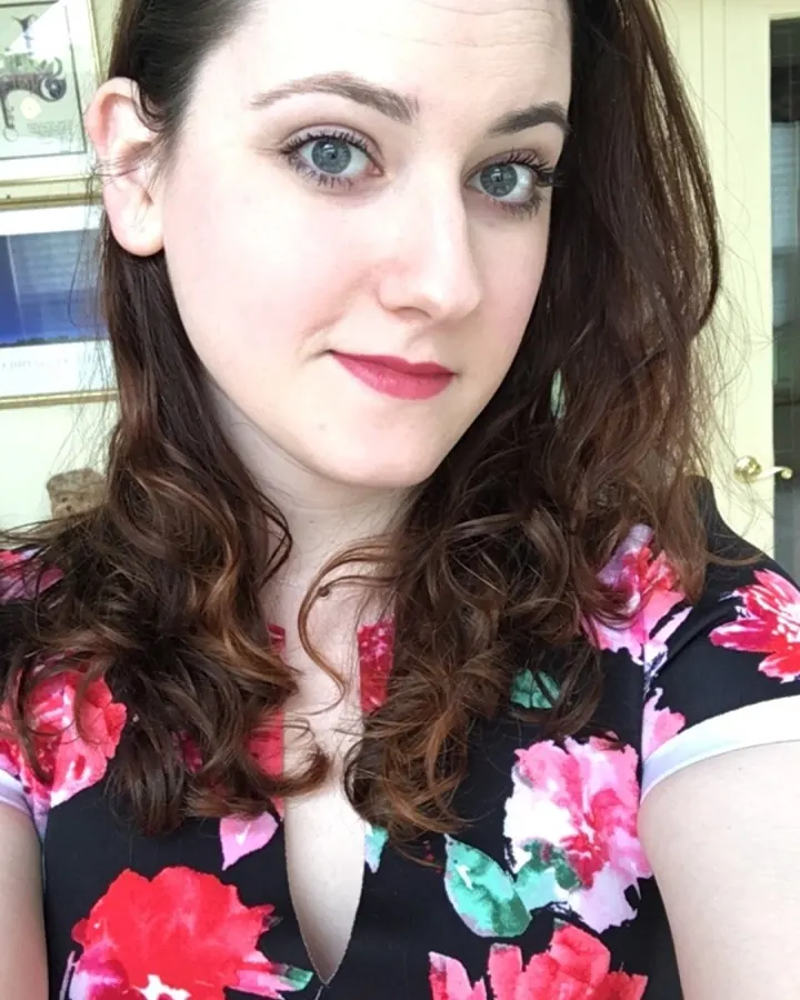 Femme aux cheveux bruns, en robe fleurie noire et rose, posant à l'intérieur avec une expression douce.
