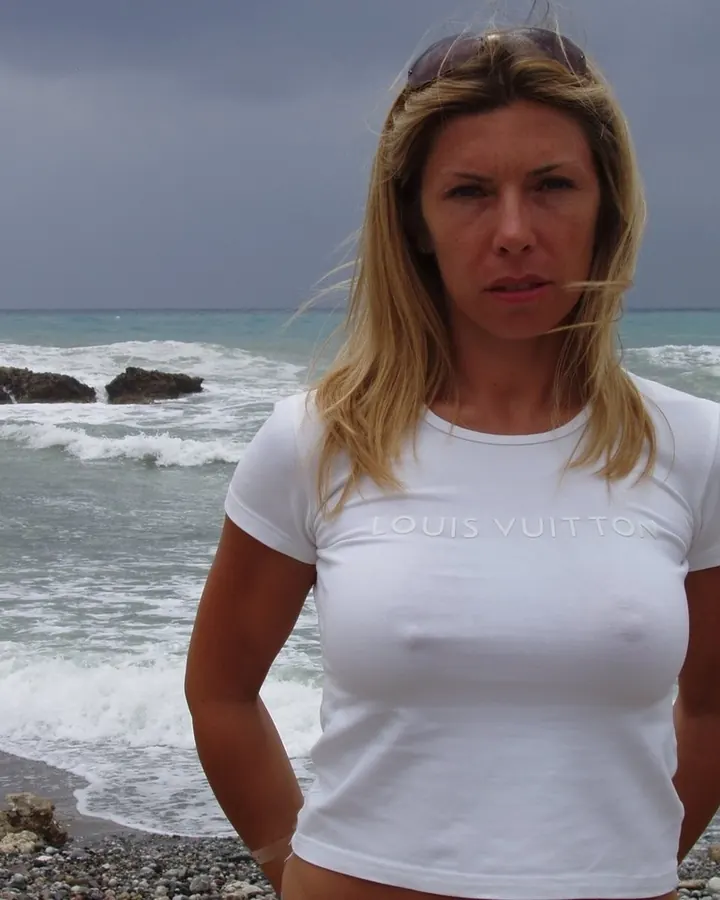 Femme aux cheveux blonds, en t-shirt blanc Louis Vuitton, posant sur une plage rocheuse avec la mer en arrière-plan.