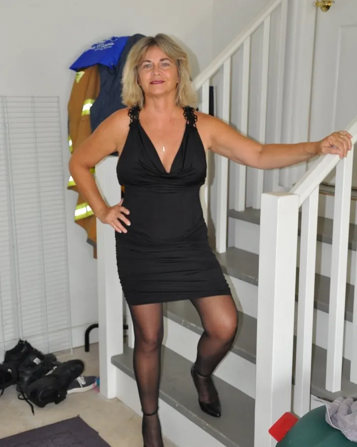 Femme blonde en robe noire, debout sur un escalier blanc, tenant la rampe, regardant l'appareil photo.
