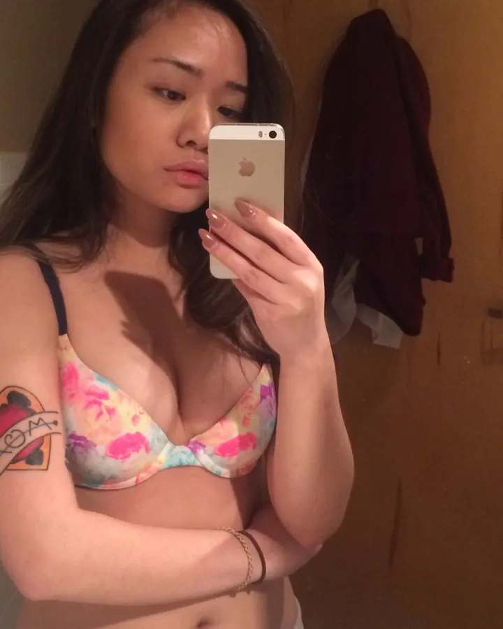 Femme asiatique en soutien-gorge coloré, prenant un selfie dans une salle de bain, tatouage visible sur le bras.