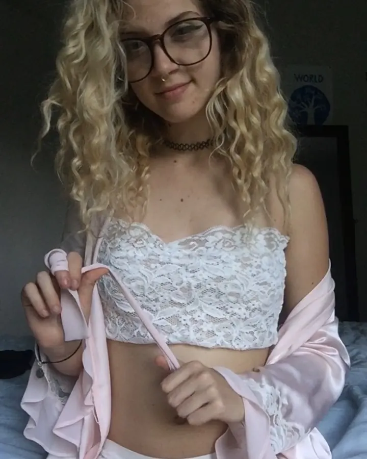 Femme blonde en lingerie blanche, lunettes, chemise rose, souriant dans un lit.
