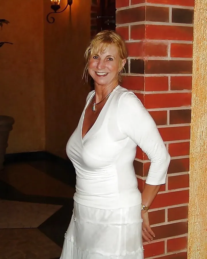 Femme blonde en robe blanche, souriant à l'appareil photo près d'un mur en briques.