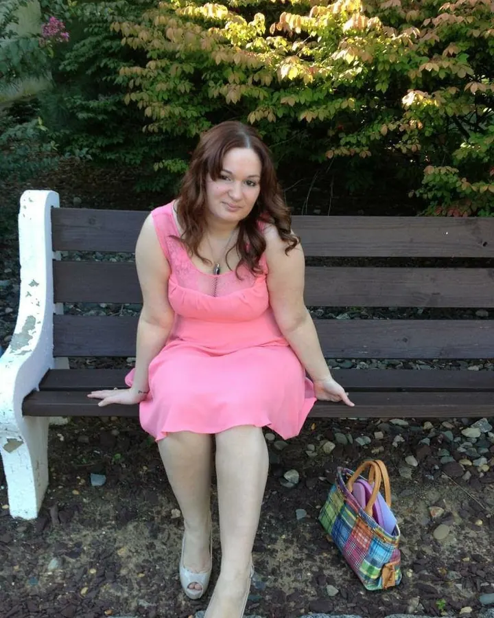 Femme brune en robe rose, assise sur un banc en bois, souriant à l'appareil photo.