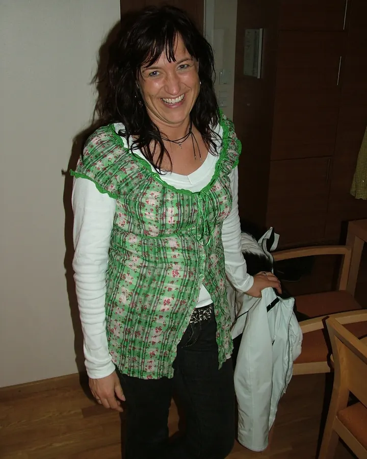 Femme brune en haut vert à fleurs, pantalon noir, souriant à l'appareil photo dans une pièce.