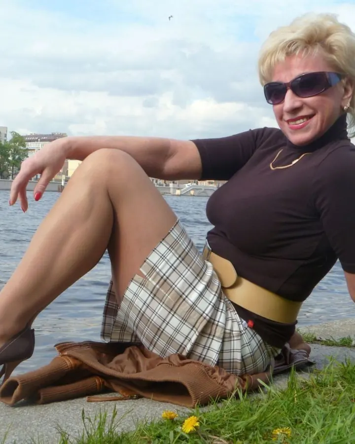 Femme d’âge mûr aux cheveux blonds courts, souriante, portant une robe blanche à motifs noirs, debout devant une cheminée en pierre.