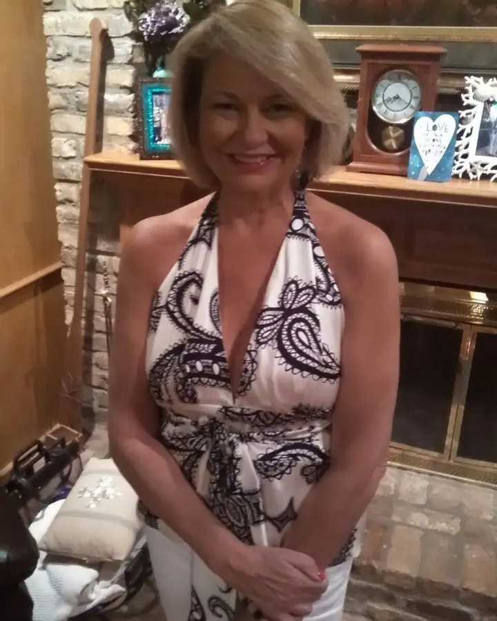 Femme d’âge mûr aux cheveux blonds courts, souriante, portant une robe blanche à motifs noirs, debout devant une cheminée en pierre.