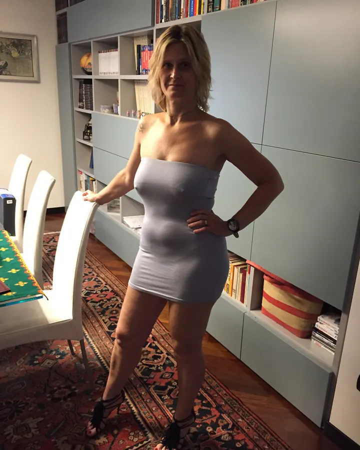 Femme aux cheveux blonds, en robe grise ajustée, chaussures à talons, debout dans un salon avec bibliothèque.