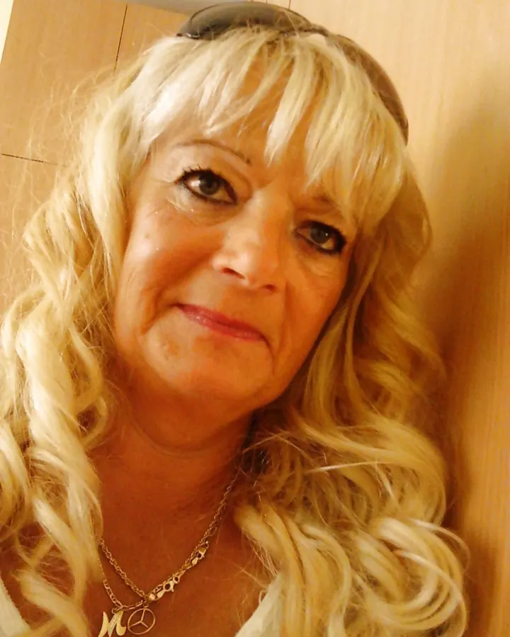 Femme aux cheveux blonds bouclés, regardant l'appareil, lunettes de soleil posées sur la tête, collier avec pendentif.