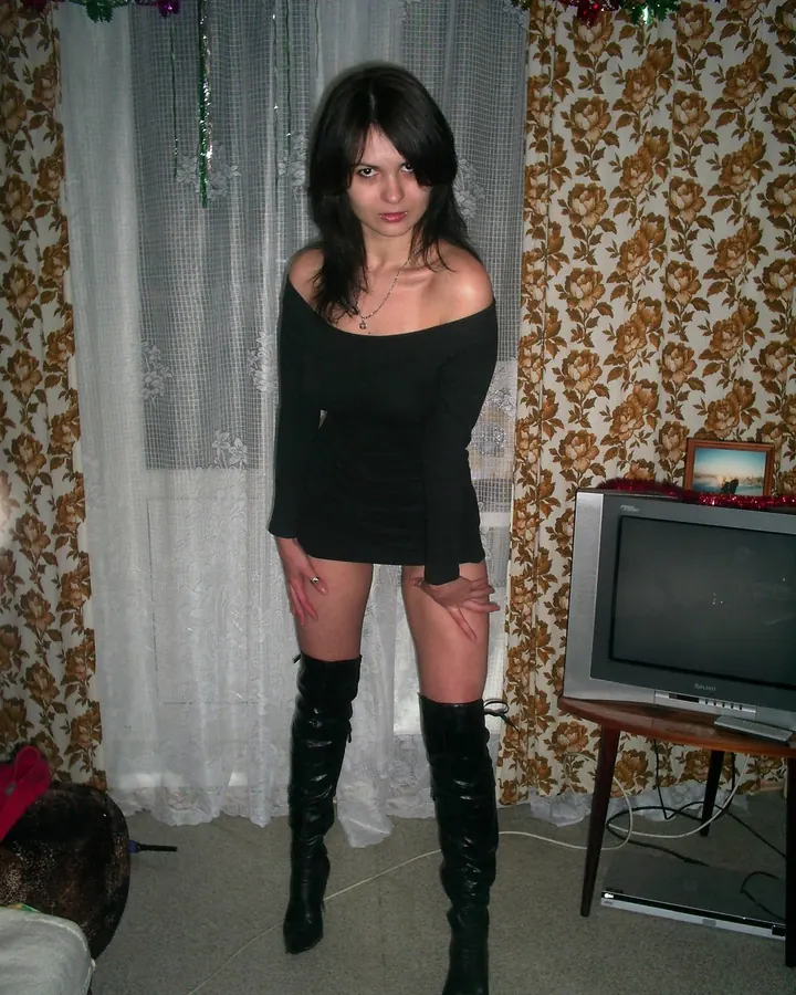 Femme aux cheveux noirs, en robe noire moulante, bottes noires, devant un rideau fleuri et un vieux téléviseur.